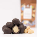 Macadamia în ciocolată fără zahăr BIO, 50g — macadamia întreagă în ciocolată, una tăiată ca să se vadă miezul, și macadamia crudă, Biovicta